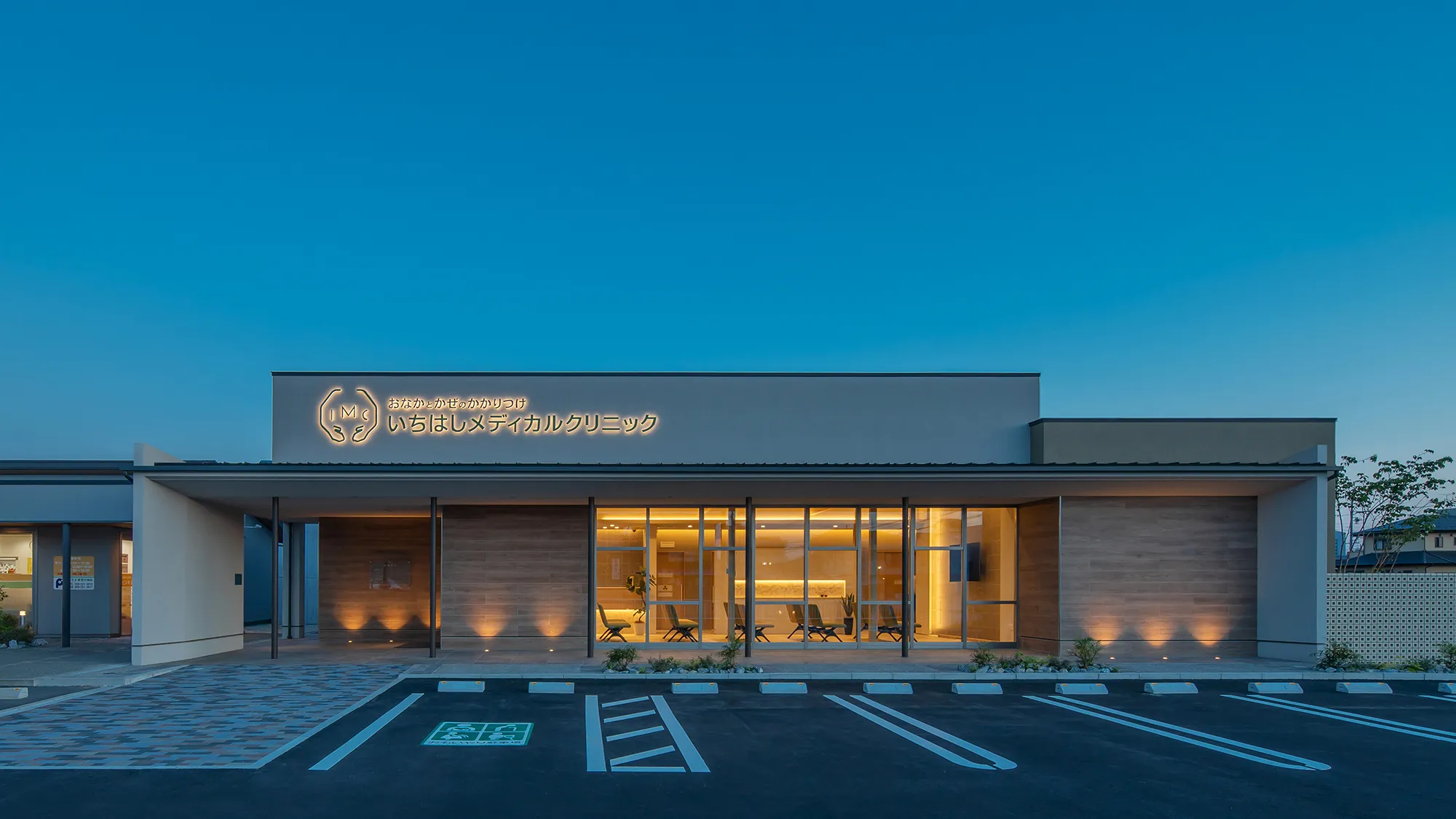 Modern medical clinic exterior at dusk with a lit sign in Japanese and a glass entrance lobby visible inside