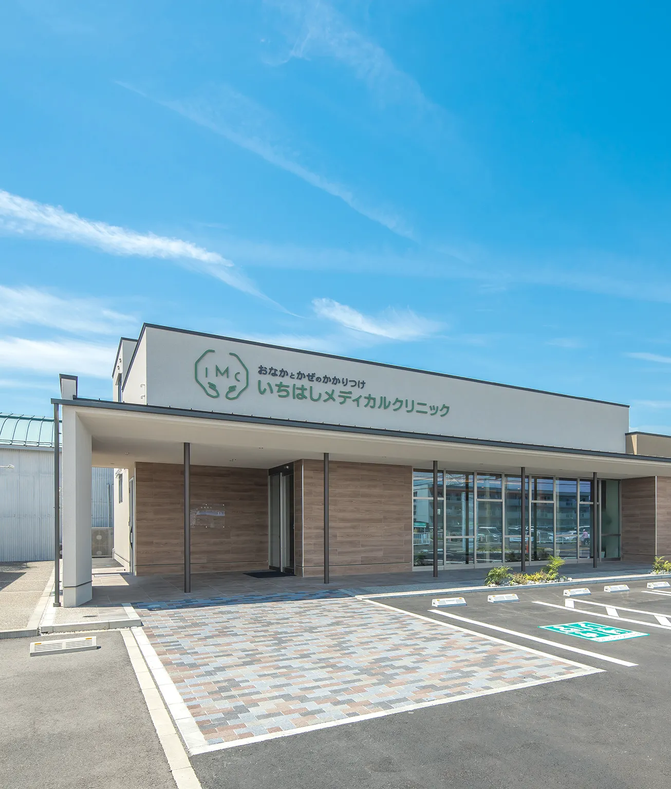 Front of a modern medical clinic with a green IMC logo and Japanese signage, glass storefront, and a tiled pedestrian walkway in front.