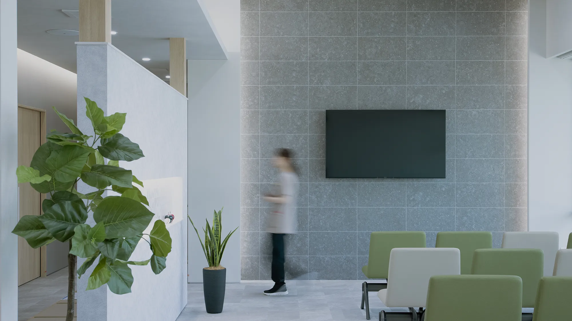 Modern lobby with a gray tiled wall, wall-mounted TV, and green/pale chairs; a blurred person walks by amid plants.