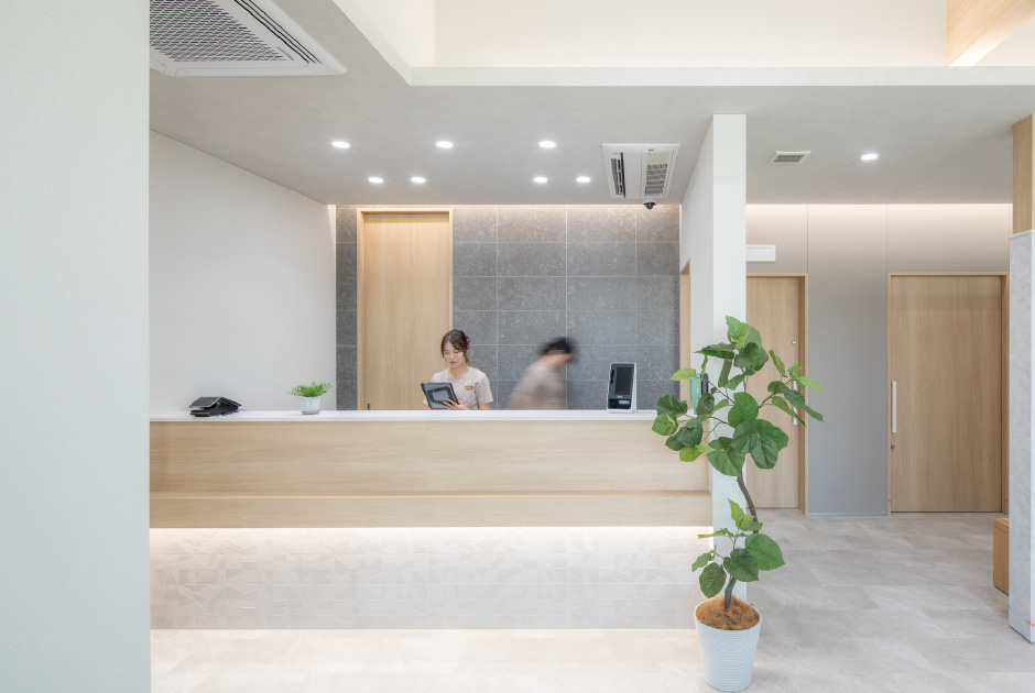 Reception area with a wooden counter and a staff member working on a tablet; potted plant nearby in a bright lobby.