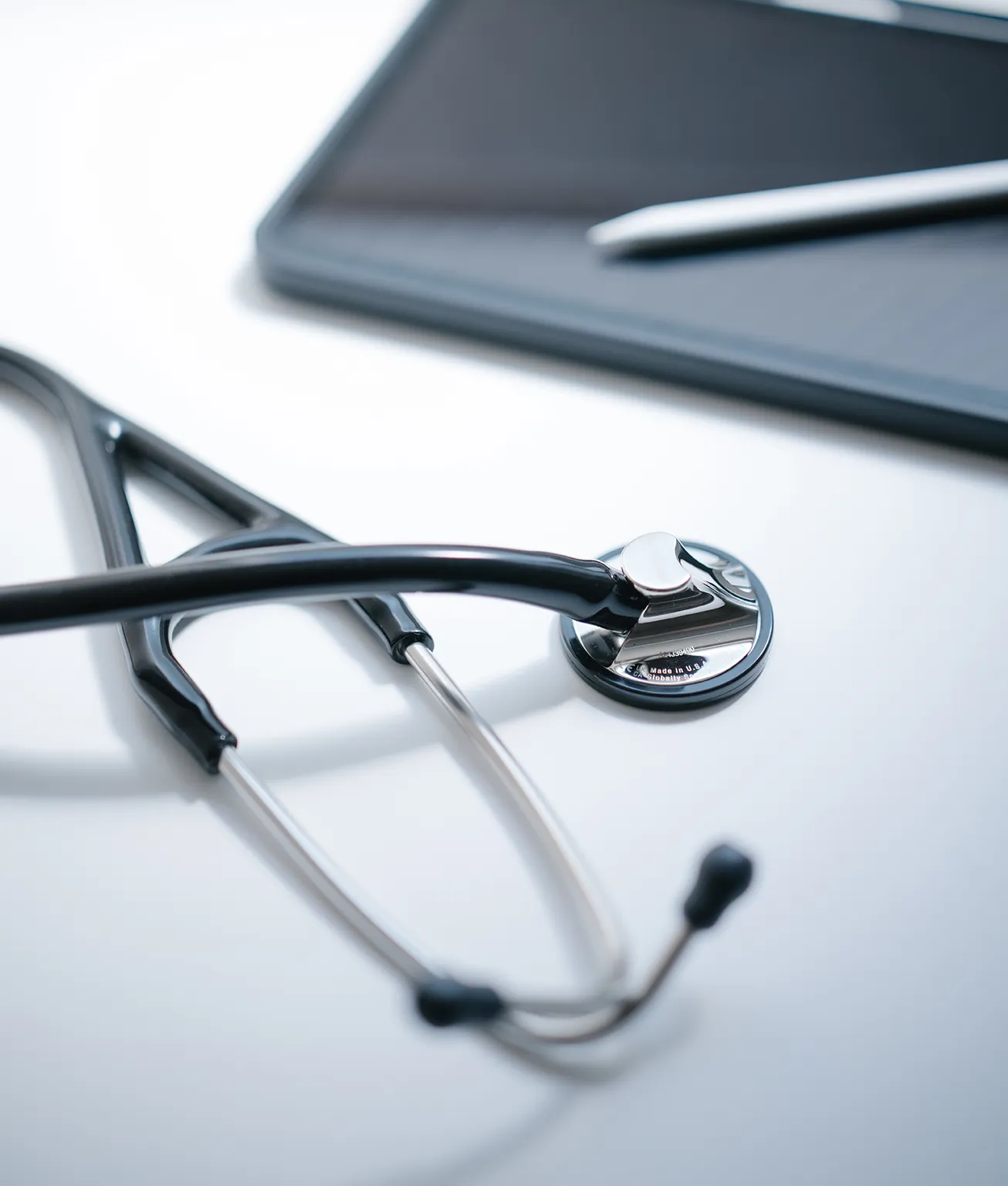 Close-up of a stethoscope resting on a white clinical surface with a blurred tablet in the background for scale and context.