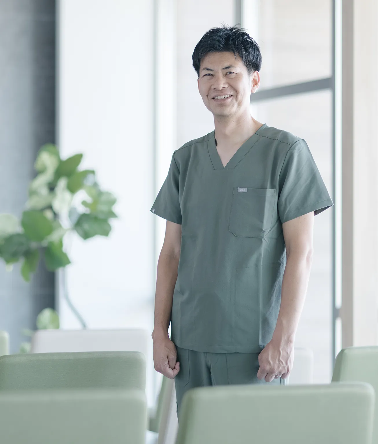 Smiling healthcare professional in green scrubs standing in a bright clinic lounge with green chairs and plants in the background.